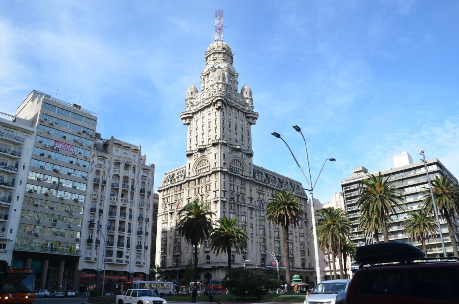 Palácio Salvo em Montevidéu, uma construção antiga e imponenete no centro da cidade, com uma torre central muito alta, ao redor, é possível ver prédios comerciais mais atuais e algumas árvores