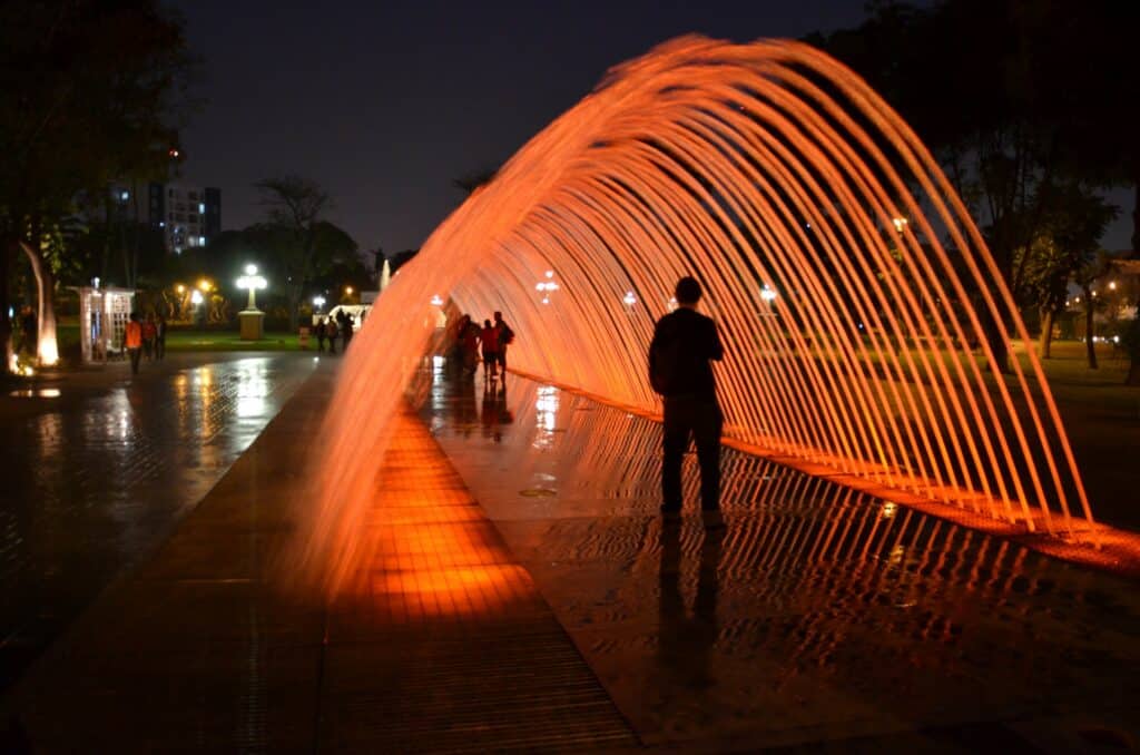 Parque de la Reserva, um local conhecida por atrações que envolvem água e luzes, na imagem é possível ver um arco se formando com esguichos de água sob uma luz alaranjada, as pessoas passam por dentro desse arco, é de noite, para representar o seguro viagem Lima