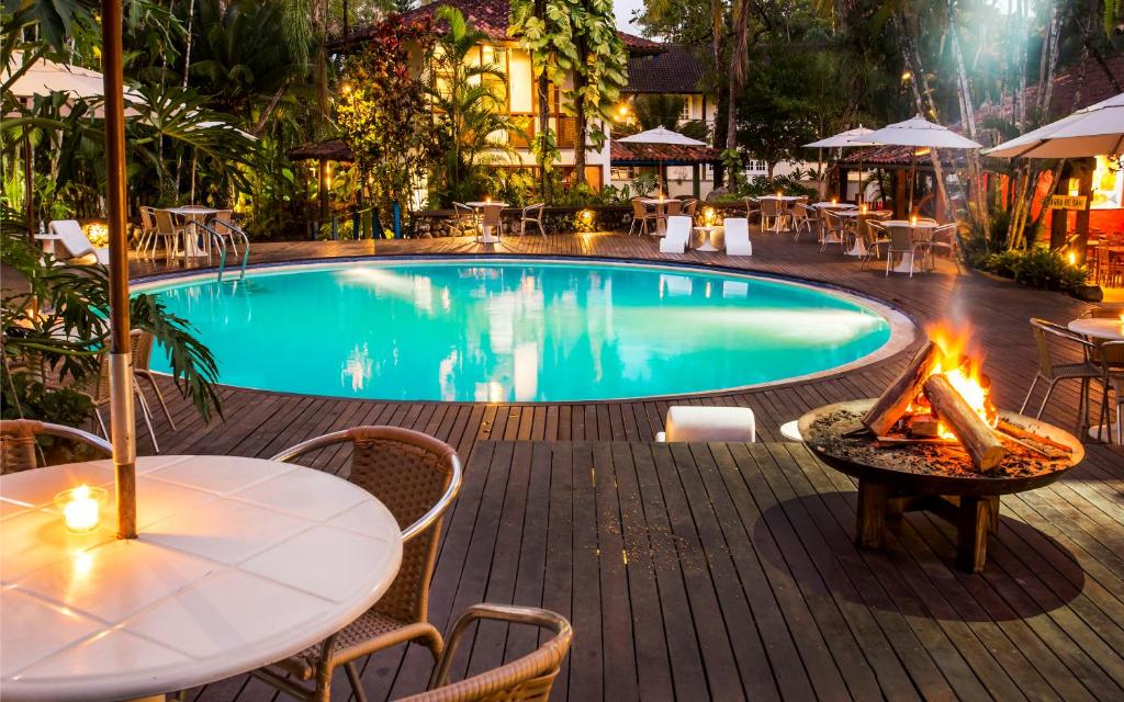 Área da piscina em Hotel Aldeia de Sahy, uma piscina redonda, com um extenso deck ao redor, com cadeiras, mesas, iluminação indireta, muitas árvores e até uma pequena fogueira, para representar pousadas em São Sebastião