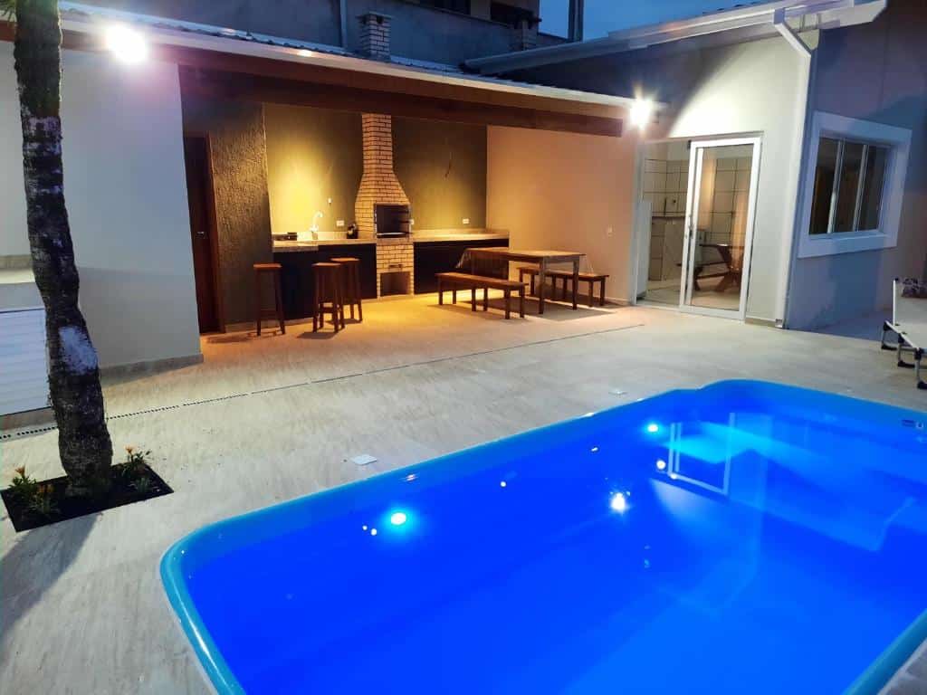 Piscina e área de churrasqueira da Vila Benatti, uma piscina pequena, uma churrasqueira com mesas e bancos de madeira e uma árvore