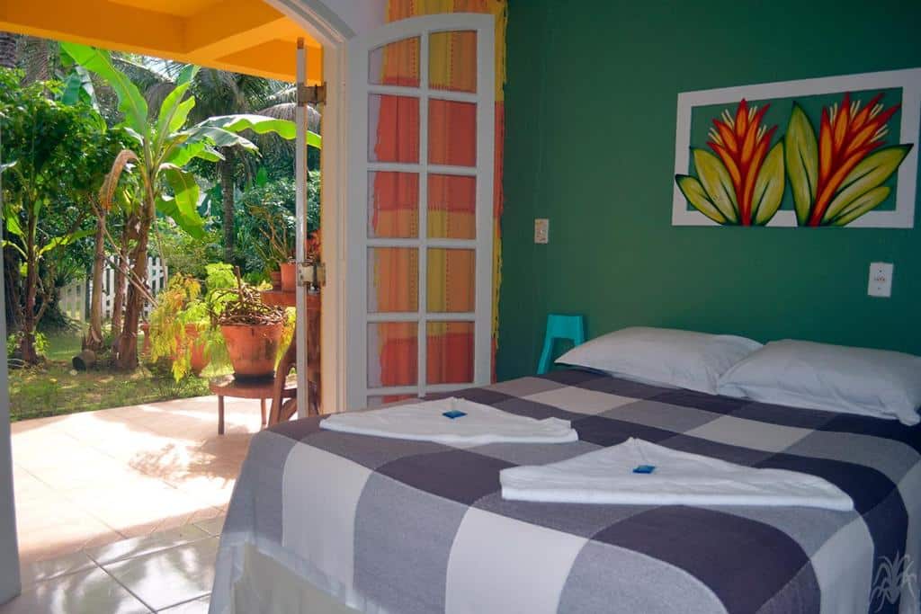 Quarto em Pousada Capim Melado, uma cama de casal ao lado de uma ampla varanda arejada e com um jardim na frente, tudo em tons de laranja e verde