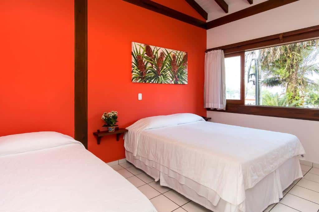 Quarto em Pousada Peixes do Mar, uma parede vermelha, uma cama de casal, um quadro e uma janela ampla do lado direito com vista para as árvores