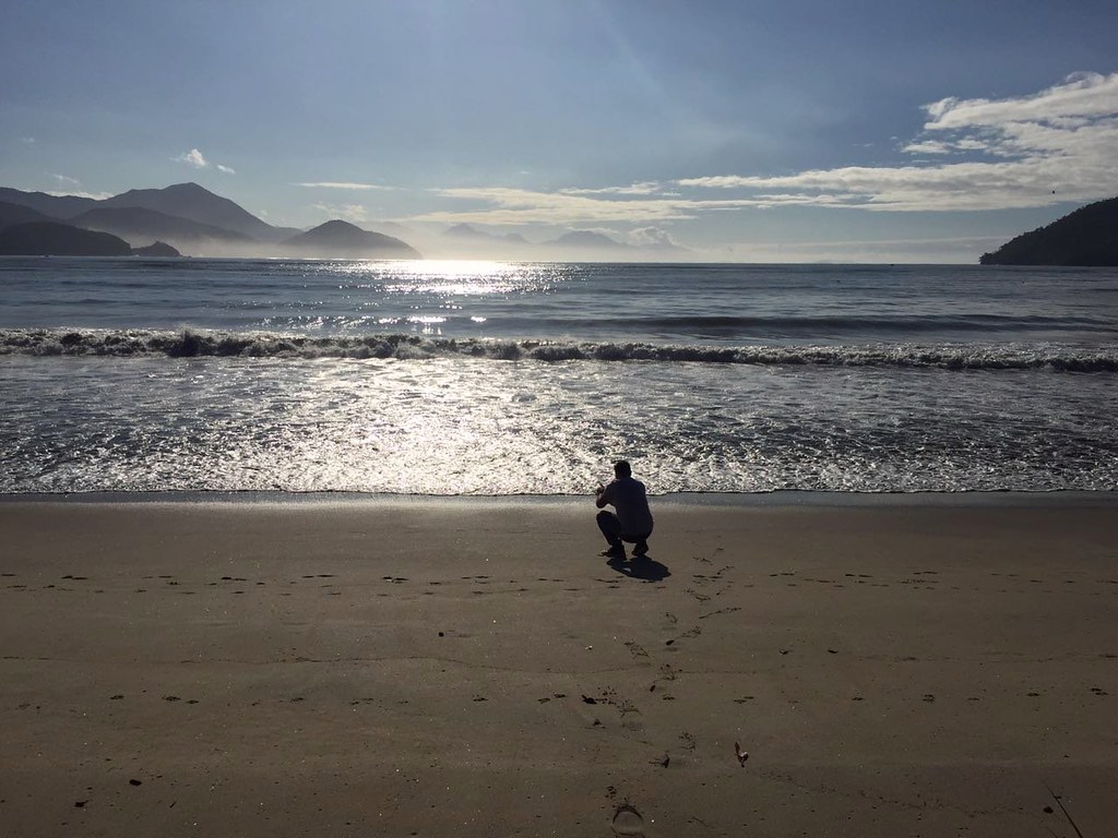 Um homem agachado próximo do mar, numa praia praticamente vazia, muitas montanhas no horizonte