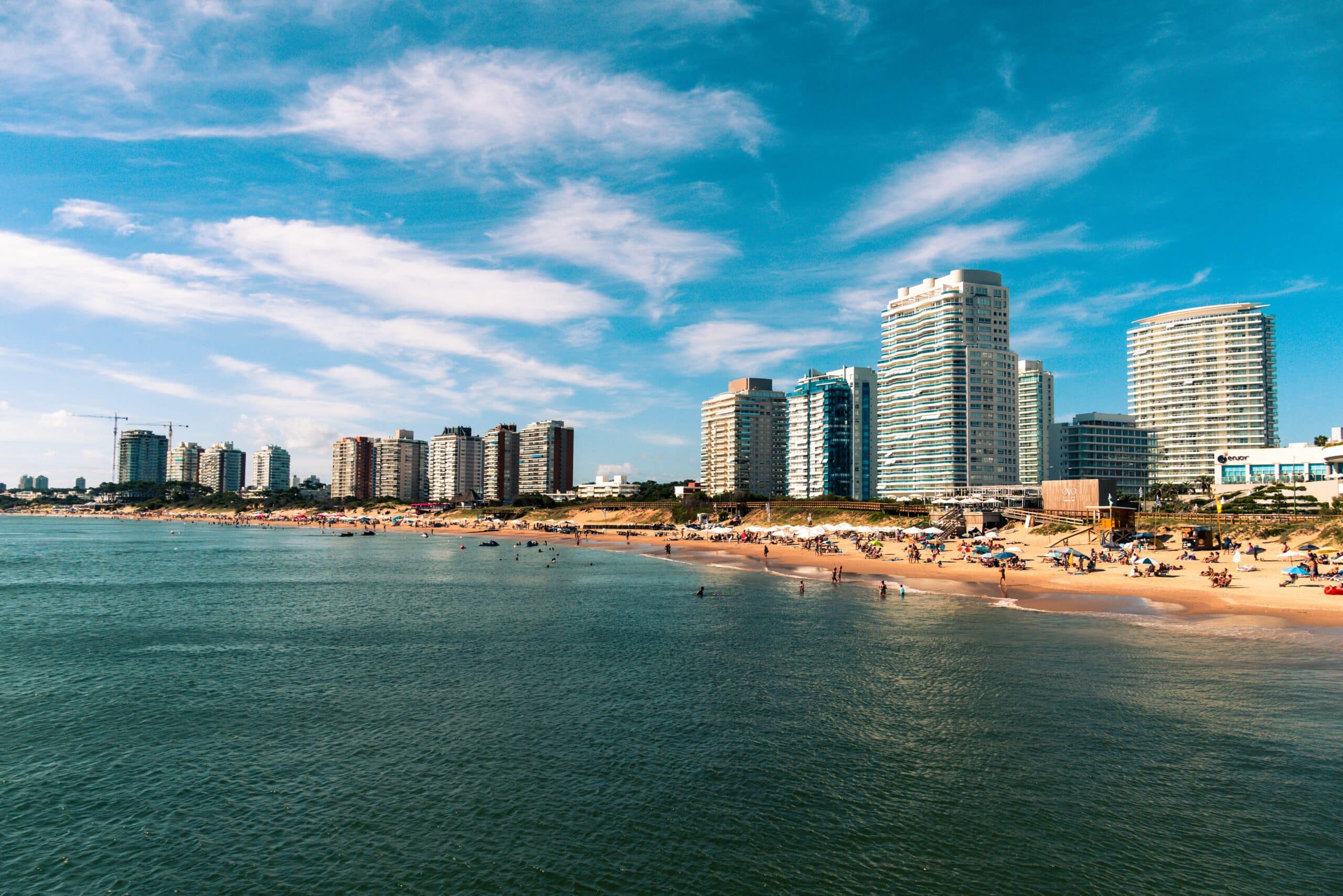 Praia de mar azul com pessoas se divertindo na areia e muitos prédios luxuosos ao fundo em um dia de céu azul ensolarado. Imagem representando um texto sobre seguro viagem Punta Del Este.