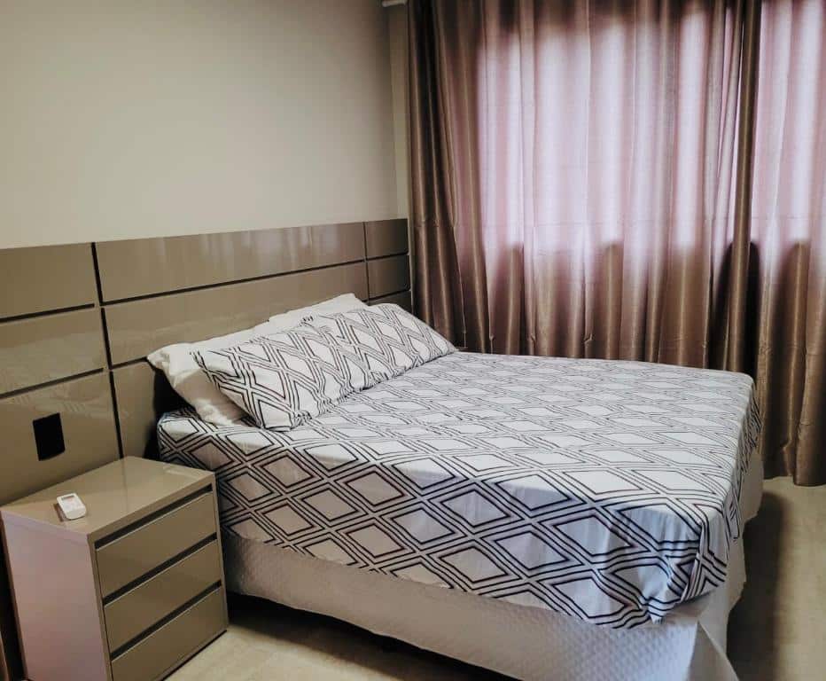 Quarto em Apartamento Alto Padrão Ubatuba, uma cama de casal, uma janela com cortinas, uma mesinha com três gavetas ao lado da cama, para representar o airbnb na Praia do Lázaro