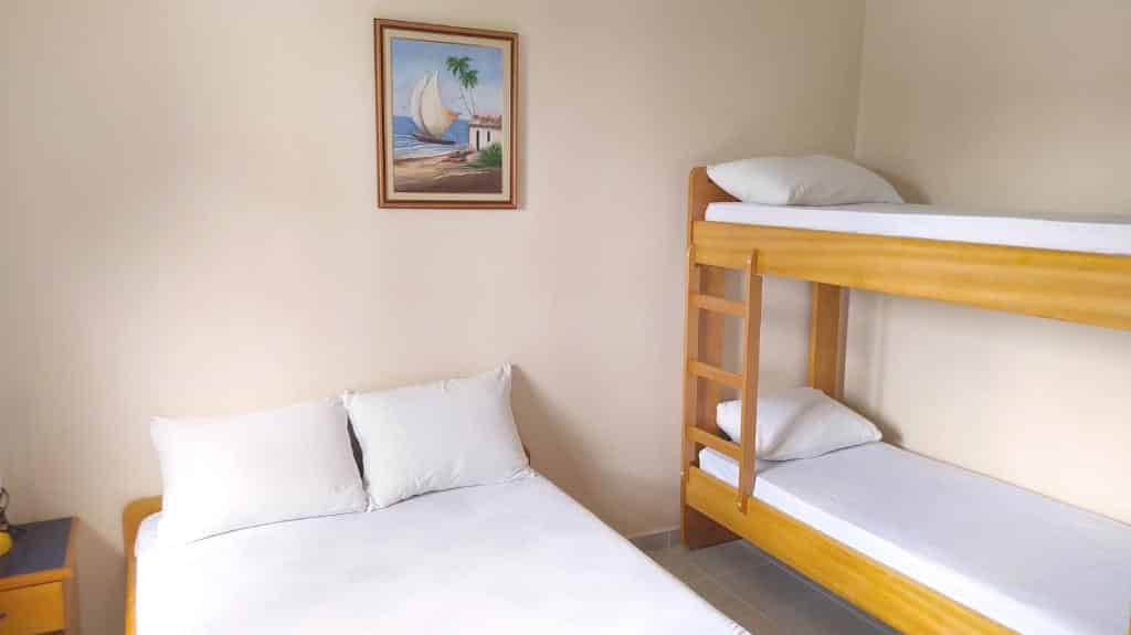 Quarto no Apartamento no Condomínio Wembley Tênis, uma cama de casal e uma beliche, roupas de cama branca e um quadro na parede central, para representar airbnb na Praia das Toninhas em Ubatuba