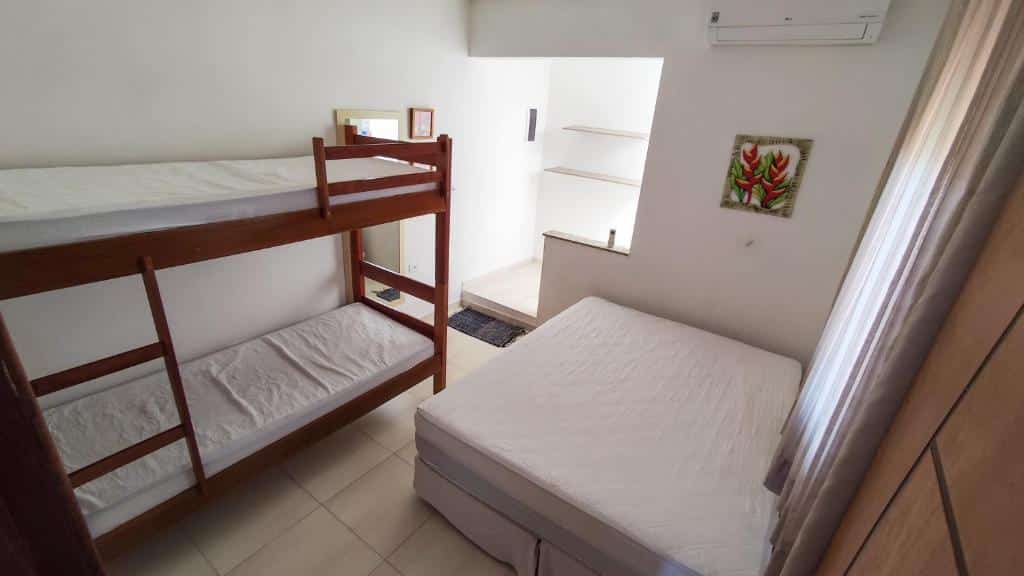 Quarto pequeno da Barreto Hospedagem com beliche, cama de casal, paredes brancas, piso de azulejo, ar-condicionado e arte na parede. Essa é uma das dicas de pousadas na Praia de Tabatinga.