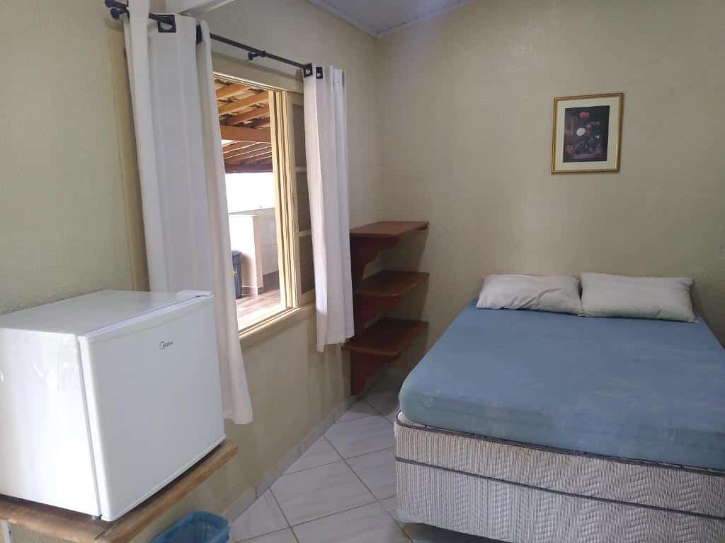 Quarto em Canto Sul Suítes, uma cama de casal, uma pequeno frigobar, uma janela ampla e algumas prateleiras