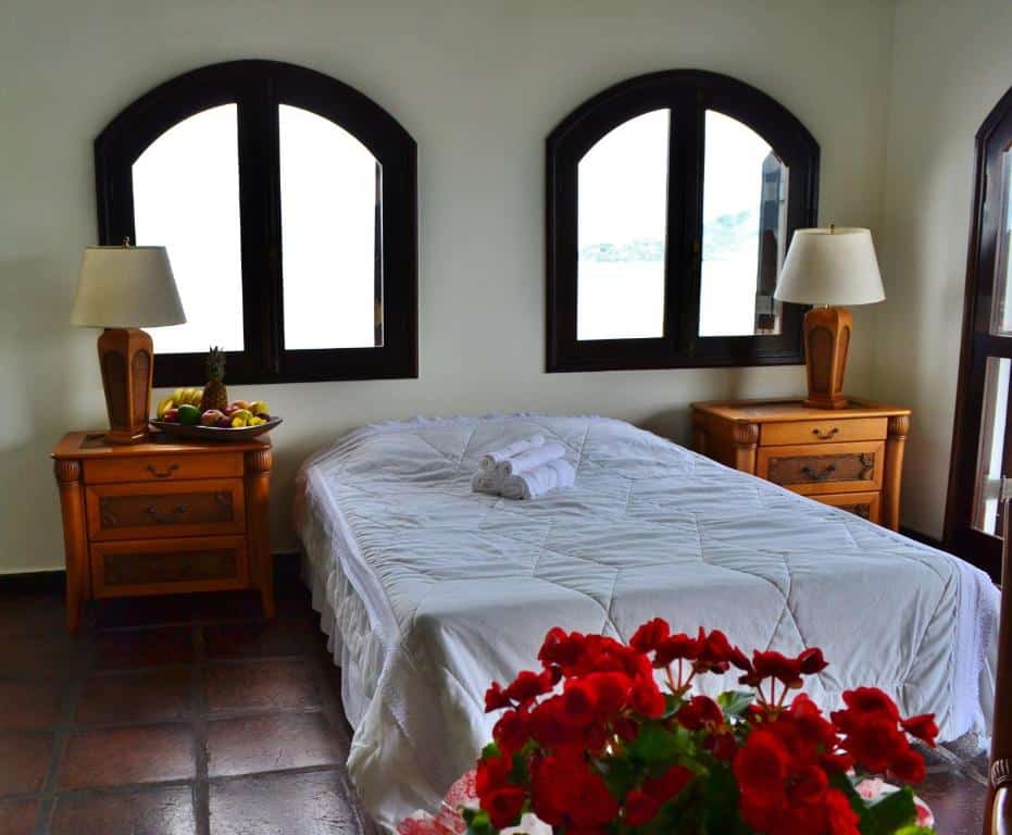 Quarto na Casa Genoveva, uma cama de casal, duas pequenas cômodas de madeira, abajures, duas janelas e uma sacada, um vaso com rosas, roupas de cama brancas, para representar airbnb em Ubatuba