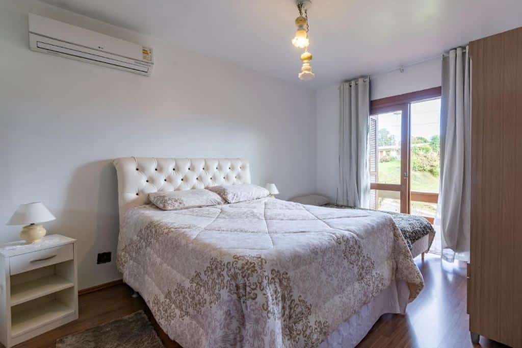 Quarto na Rosa Casa Tirol Gramado, uma sacada com cortinas, uma cama de casal, uma mesinha com um abajur, um ar-condicionado e um armário, para representar airbnb em Gramado