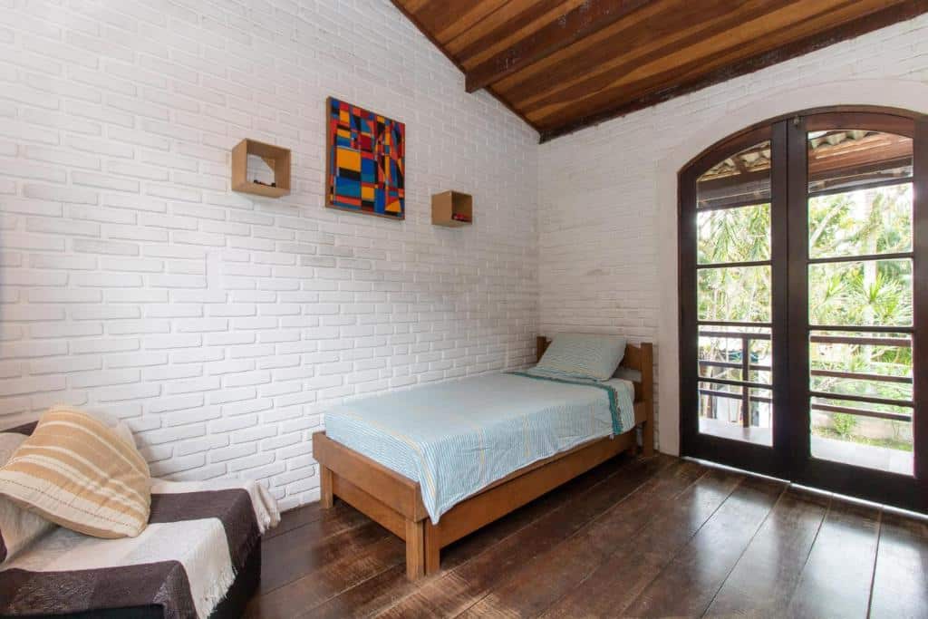 Quarto na Casa Tranquila, chão de madeira, uma sacada, parede de alvenaria em tijolinhos brancos, uma poltrona com uma almofada e um quadro colorido na parede