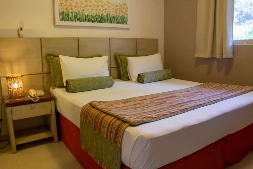 Quarto em Ciribaí Praia Hotel, uma cama de casal, uma cômoda ao lado com uma luminária e um telefone, uma janela ao lado e um quadro sob a cama