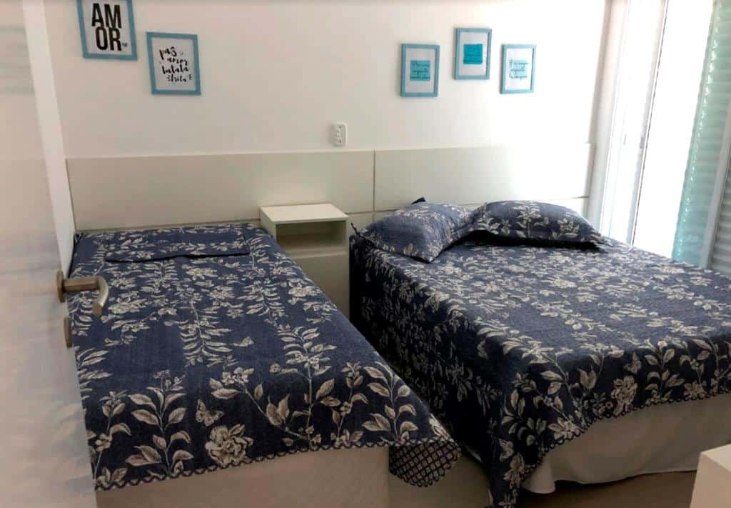 Quarto na Cobertura Alto Padrão Vista para o Mar, uma cama de casal e uma de solteiro, uma sacada, roupa de cama e travesseiros azuis com desenho de flores, para representar airbnb em Ubatuba