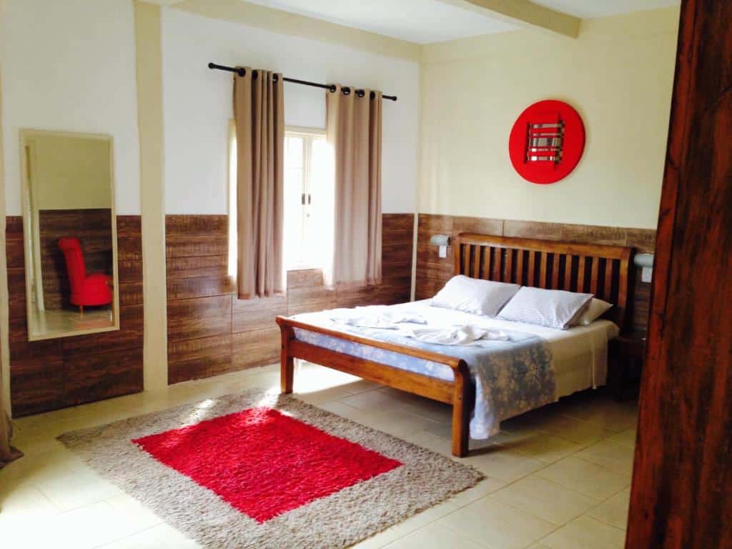 Quarto na Pousada Ananda-ri, local amplo, com uma cama de casal, uma janela, um espelho e um tapete, os detalhes são em vermelho e bege