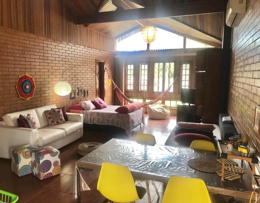 Sala de plano aberto da EccO Capricornio com paredes de tijolos, camas, sofá, rede, cadeiras amarelas, uma mesa e janelas grandes que permitem a entrada de luz. Essa é uma das dicas de pousadas na Praia de Tabatinga.