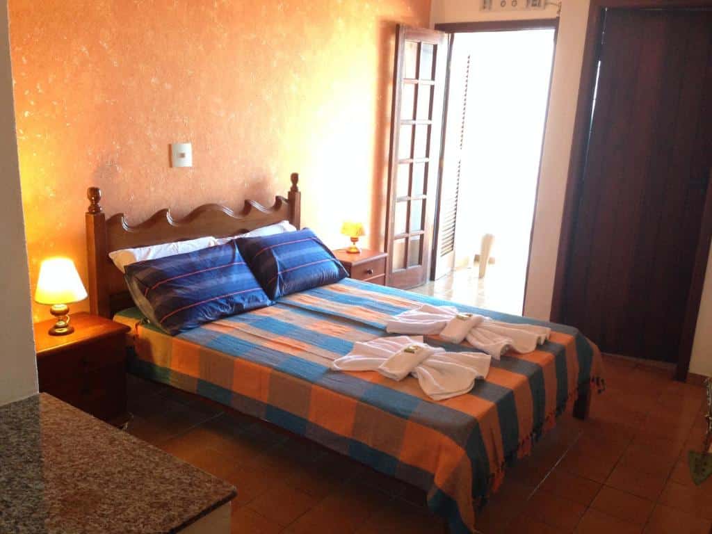 Quarto no Enseada Hotel, uma cama de casal, com toalhas e travesseiros sob a cama, uma sacada, um banheiro e um abajur ao lado da cama, para representar pousadas na Praia da Enseada em Ubatuba
