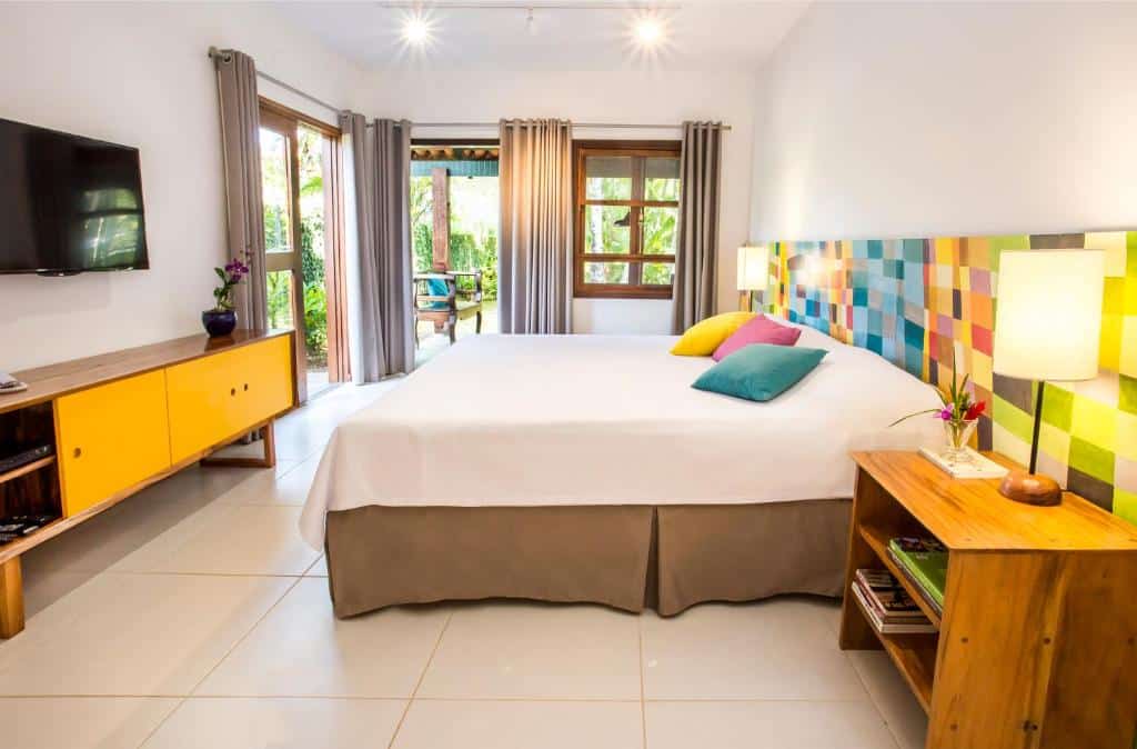 Quarto do Hotel Aldeia de Sahy, um quarto amplo com uma cama de casal, abajures, uma estante e uma televisão, uma sacada ampla com cortinas, iluminação indireta, as cores principais da decoração são o amarelo, o rosa e o amarelo