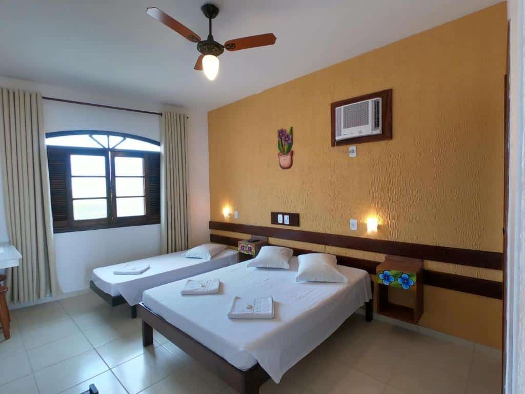 Quarto em Hotel Parque Atlântico, uma cama de casal e uma de solteiro, uma janela com cortina, almofadas e toalhas, um ar-condicionado e luzes indiretas perto da cama, para representar pousadas no Centro de Ubatuba