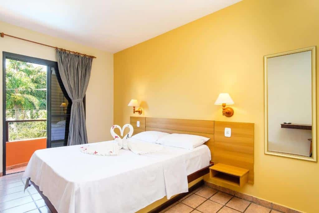 Quarto no Hotel Ponta das Toninhas, uma cama de casal, dois abajures, uma espelho e uma pequena sacada, sob a cama algumas toalhas e travesseiros, para representar a Praia das Toninhas