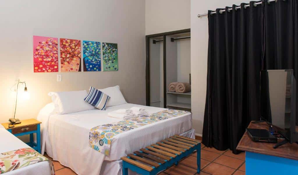 Quarto no Hotel Porto Di Mare, uma cama de casal, uma pequena mesa ao lado da cama com um abajur, alguns quadros coloridos, um pequeno armário, uma televisão e cortinas na sacada, para representar pousadas na Praia da Enseada em Ubatuba