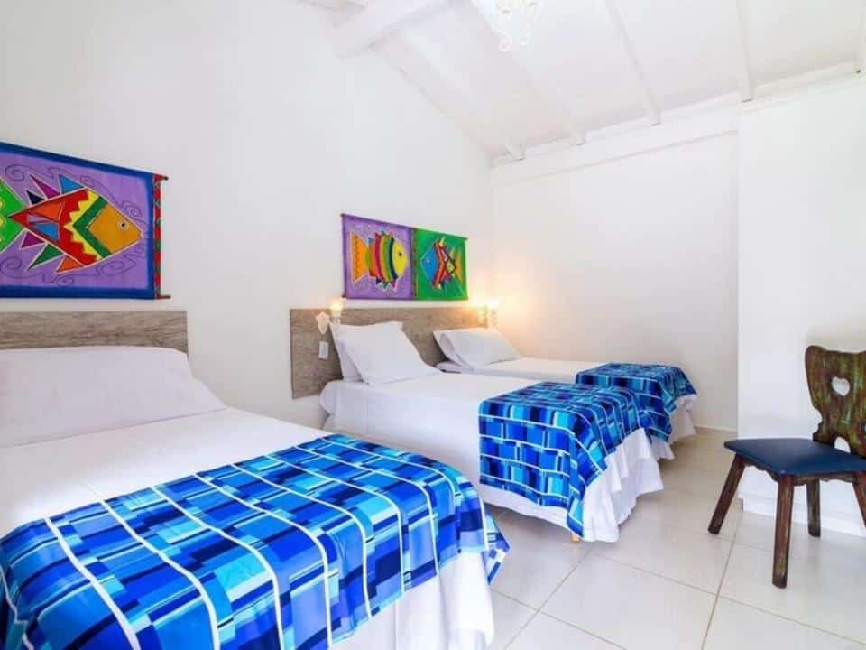 Quarto no Hotel Village Enseada, uma cama de casal e duas de solteiro, tudo em tons de branco e azul, uma cadeira rústica, alguns quadros com peixes desenhados, para representar pousadas na Praia da Enseada em Ubatuba
