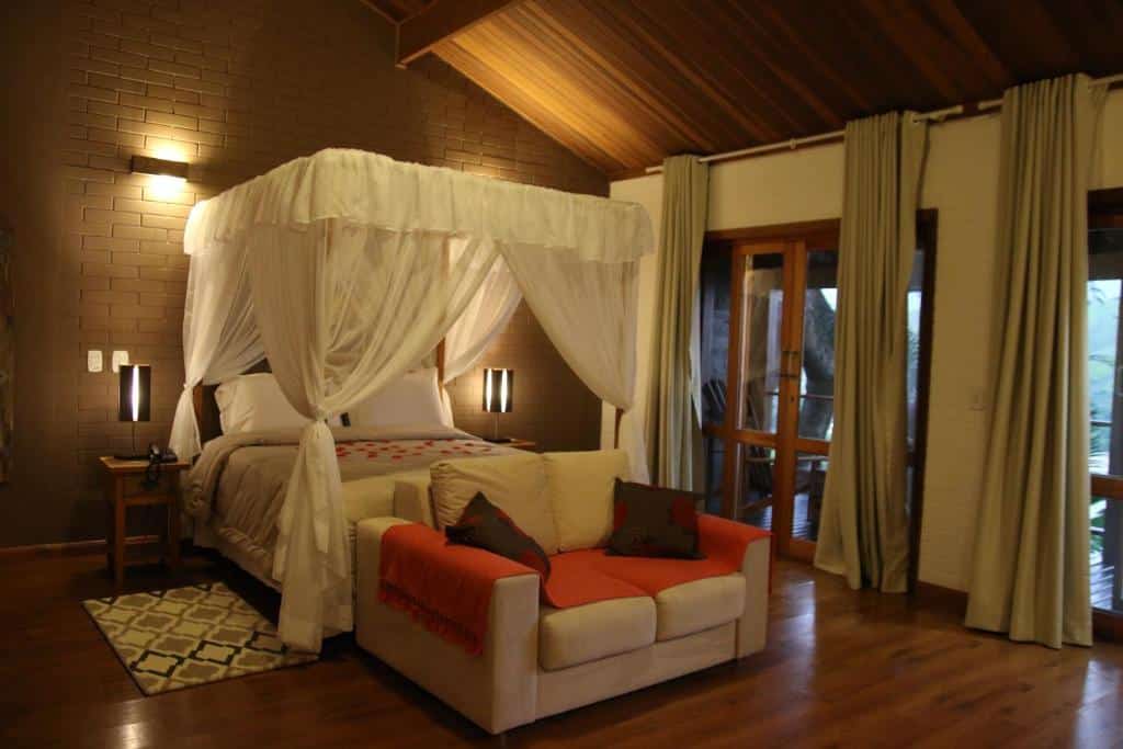 Quarto em Ilha de Toque Toque Boutique Hotel & Spa, uma cama de casal num quarto amplo, uma sacada, uma pequeno sofá, chão de madeira, abajures e tudo em tons de bege e verde