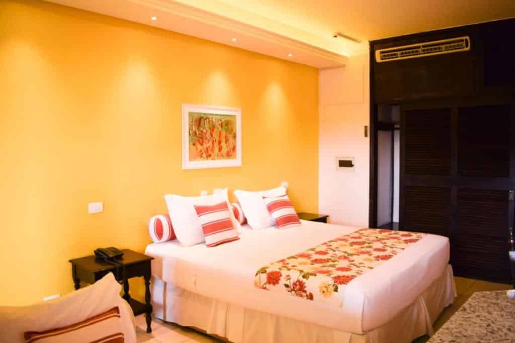 Quarto no Ubatuba Palace Hotel, uma cama de casal, decoração em tons de laranja, branco e vermelho, uma pequena mesa com um telefone, uma quadro sob a cama, ar-condicionado e um armário, para representar pousadas no Centro de Ubatuba