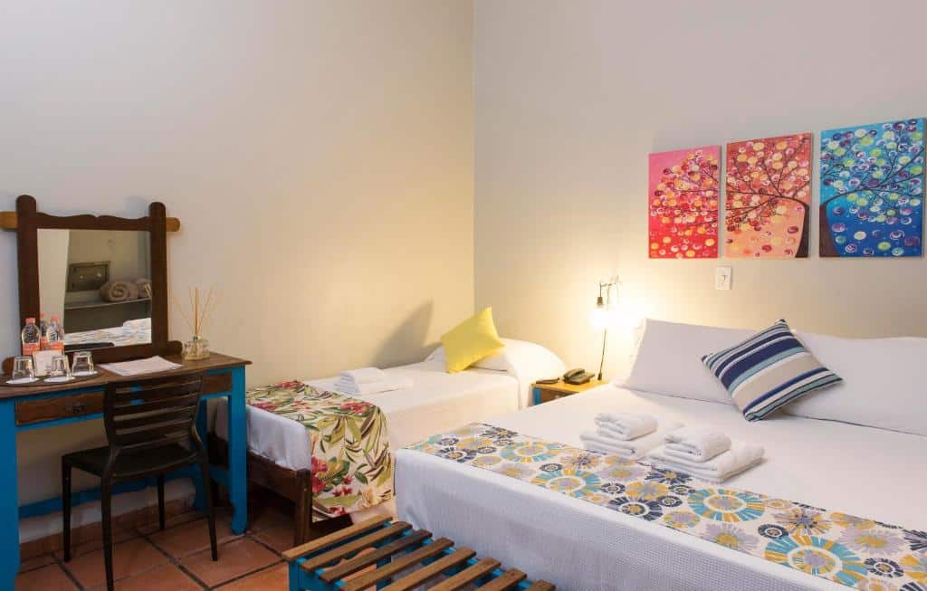 Quarto em Hotel Porto Di Mare, uma cama de casal, uma de solteiro, muitas alfomadas, travesseiros e toalhas sob as camas, decoração minimalista em tons de azul e amarelo, uma pequena penteadeira azul de madeira no canto esquerdo e três quadros sob as camas