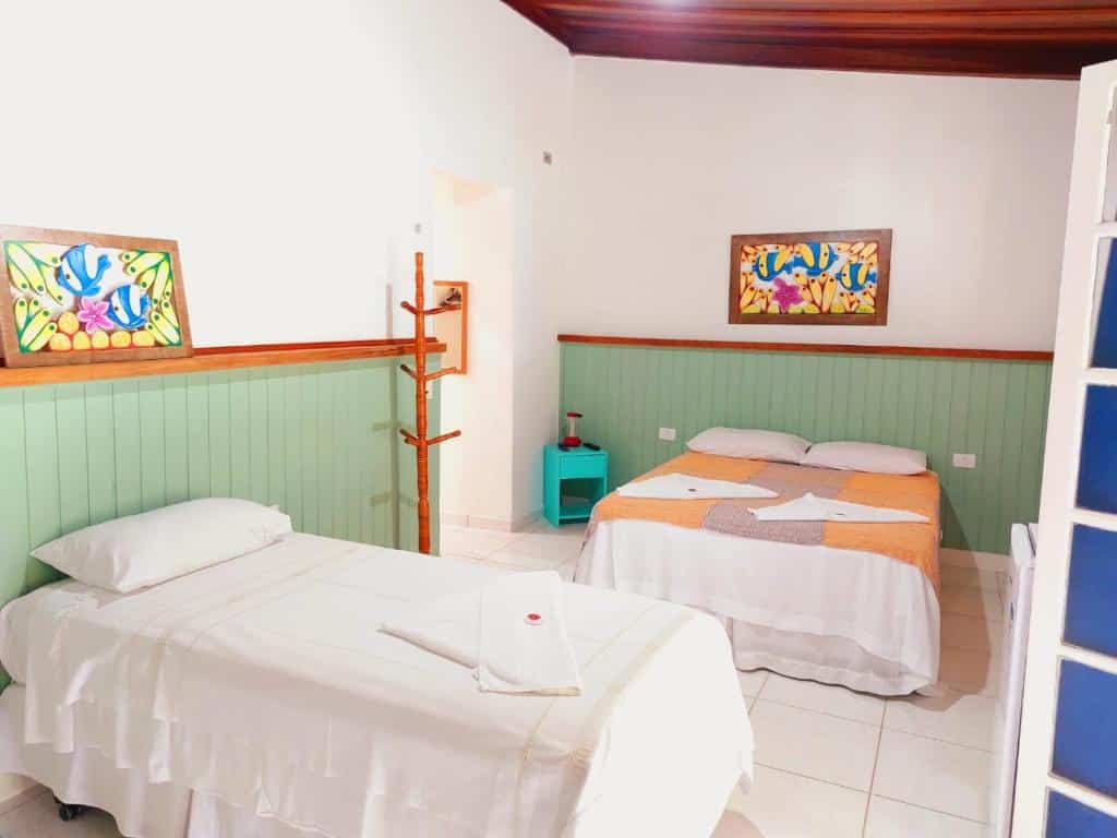 Quarto da Pousada Capim Melado, uma cama de casal e uma de solteiro, paredes em tom de verde claro, quadros coloridos, um cabideiro, um móvel azul com um abajur do lado da cama