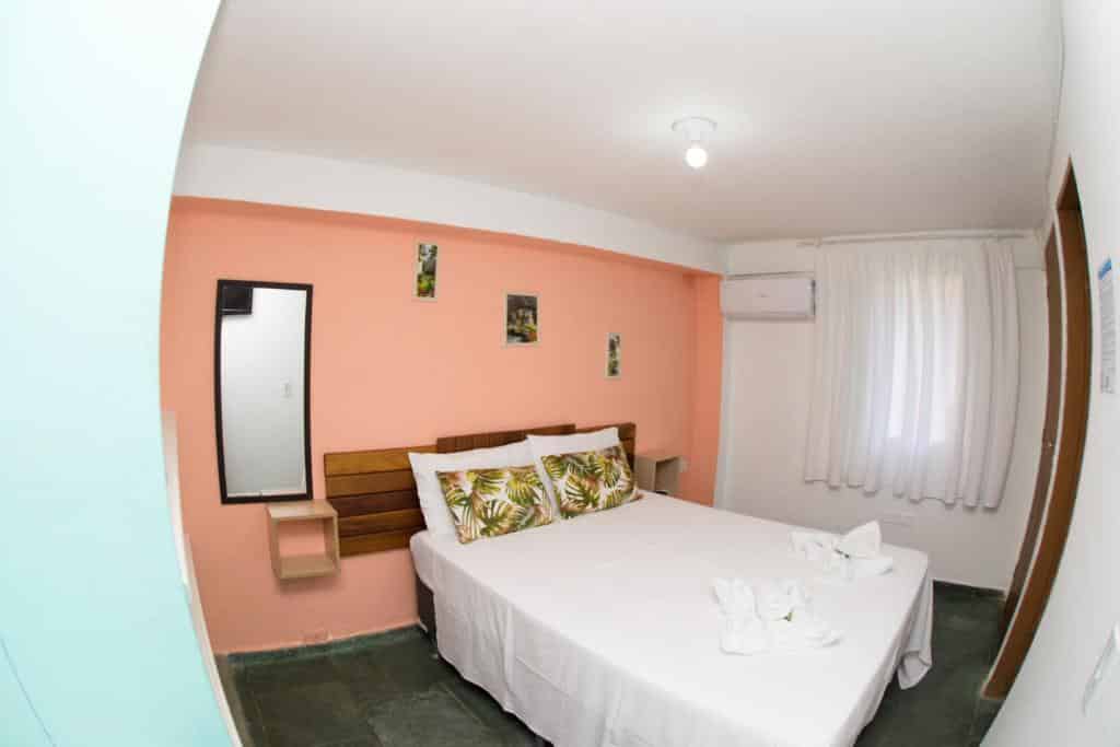 Quarto em Pousada Viva Ubatuba, uma cama de casal com travesseiros e toalhas, um espelho, um ar-condicionado, uma janela com cortinas brancas, para representar pousadas na Praia das Toninhas