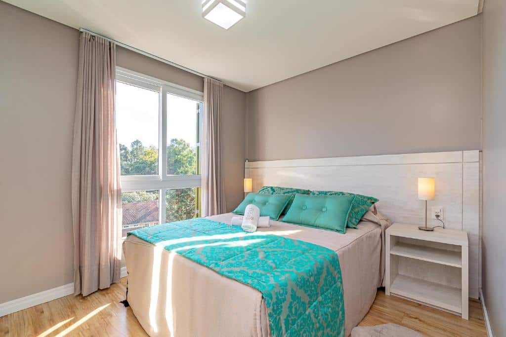 Quarto no Rosa Apartamento Giovani Pizetta, uma cama de casal, uma poltrona, uma mesa com abajur, toalhas e travesseiros sob a cama, tudo em tons de verde claro e cinza, para representar airbnb em Gramado