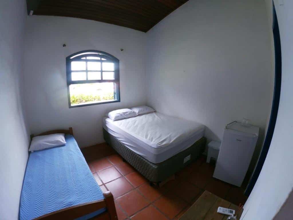 Quarto em Suítes Pé na Areia, uma cama de casal, uma de solteiro, um frigobar e uma janela, pousadas na Barra do Sahy
