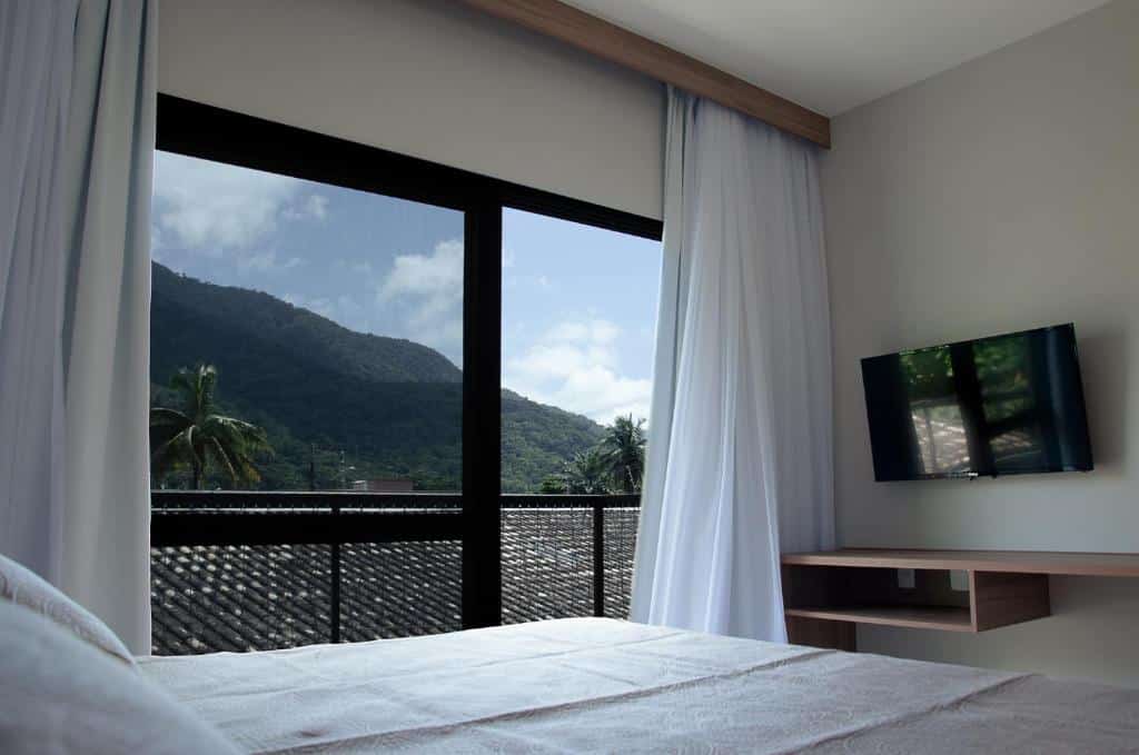 Quarto em Tons do Mar Residence na Praia do Lázaro, um quarto com uma sacada grande com vista para as montanhas, uma cama de casal e uma televisão de frente para a cama