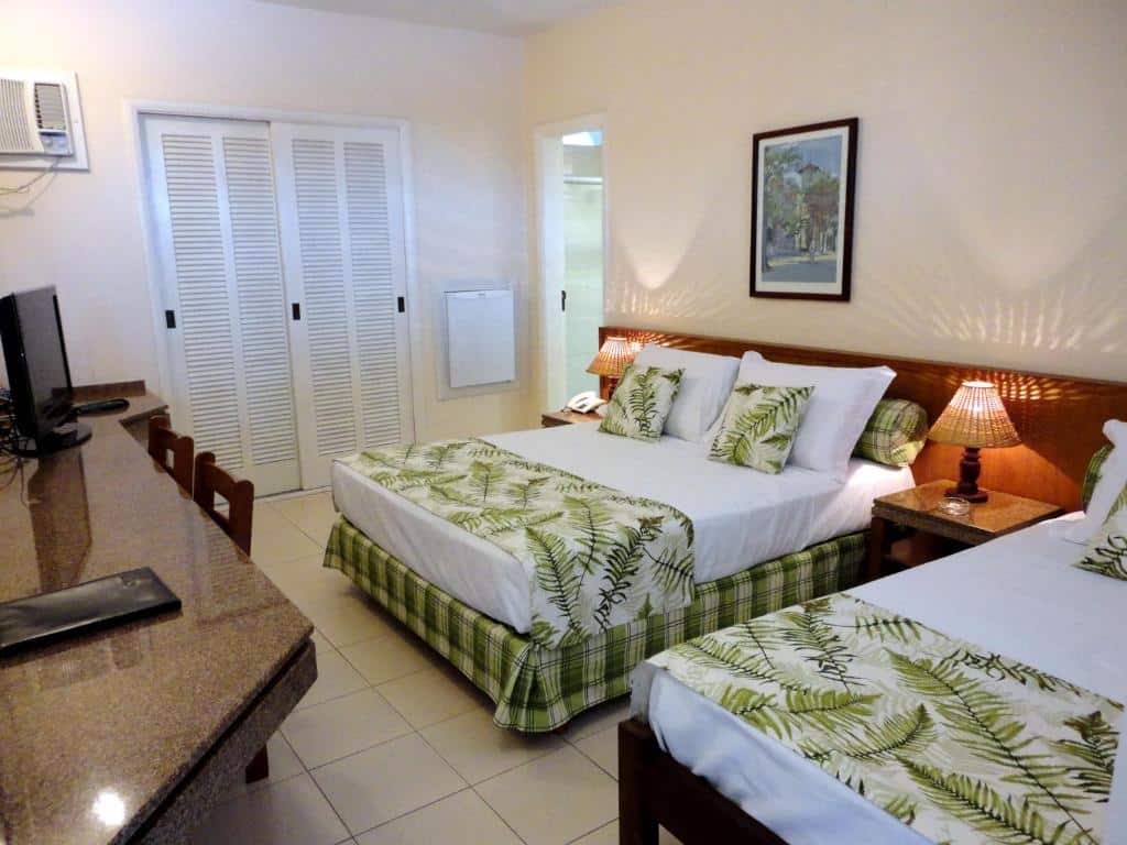 Quarto no Ubatuba Palace Hotel, uma cama de casal, um abajur, uma cama de solteiro, um armário, ar-condicionado, uma televisão, um balcão com duas cadeiras, roupas de camas com estampas tropicais