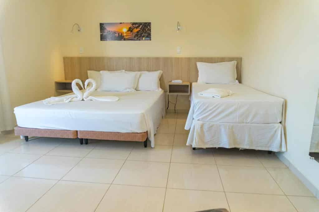 Quarto no  Ubatuba Praia Hotel, uma cama de casal, uma de solteiro, um espelho e um quadro, para representar pousadas na Praia do Tenório