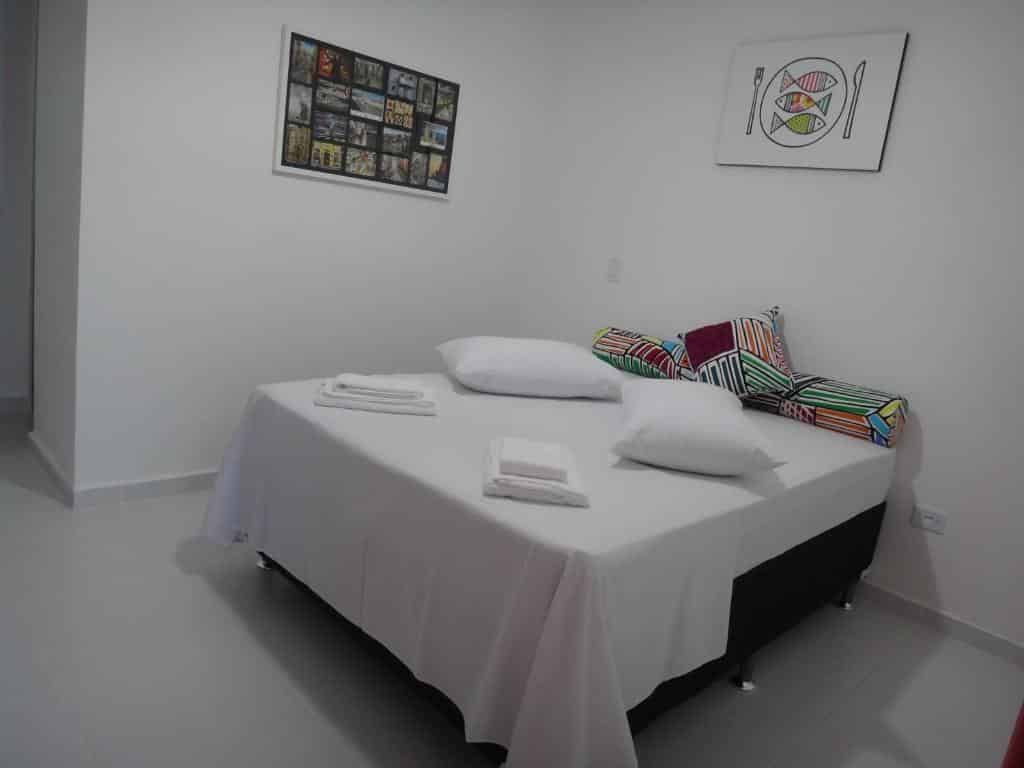 Quarto em Urbano Suites, uma cama de casal com travesseiros, almofadas e toalhas sob a cama, além de alguns quadros nas paredes, para representar pousadas no Centro de Ubatuba