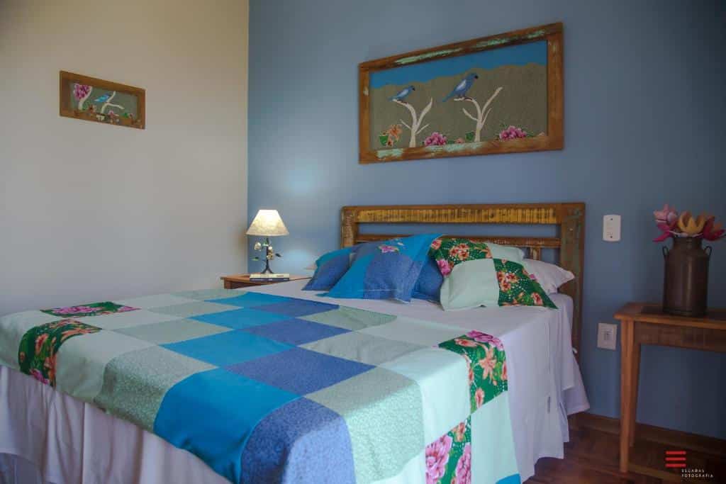 Quarto na Vila Chico, uma cama de casal com colchas de retalhos coloridos, toda a decoração com estilo de roça e detalhes em madeira, uma mesa com um abajur e dois quadros