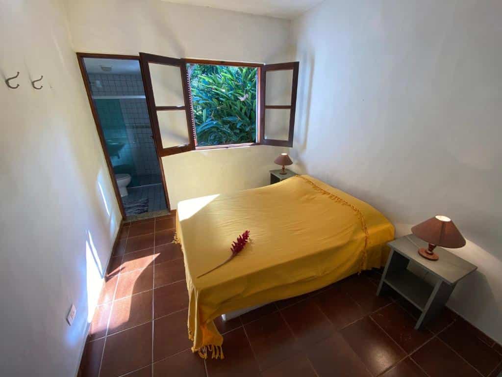Quarto em Vila dos Tangarás, uma cama de casal, dois abajures, um banheiro e uma janela que dá vista para o jardim