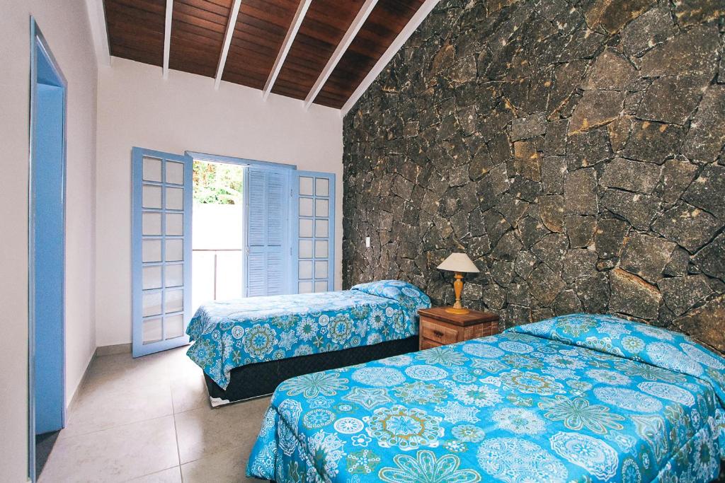 Quarto em Villa Divina Pietra - Ubatuba, duas camas de solteiro, com cobertas azuis, uma parede de pedra rústica, uma sacada, para representar airbnb na Praia das Toninhas em Ubatuba