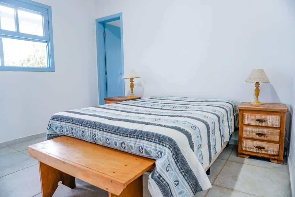 Quarto na Villa Divina Pietra, uma cama de casal amplo, móveis de madeira e dois abajures, uma janela e um banco, para representar airbnb em Ubatuba