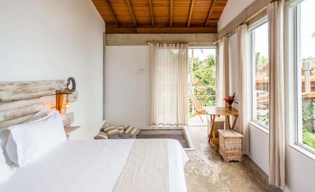 Quarto em Villa Sapê Pousada, uma cama de casal, alguns móveis rústicos de madeira, uma sacada e janelas amplas com cortinas claras