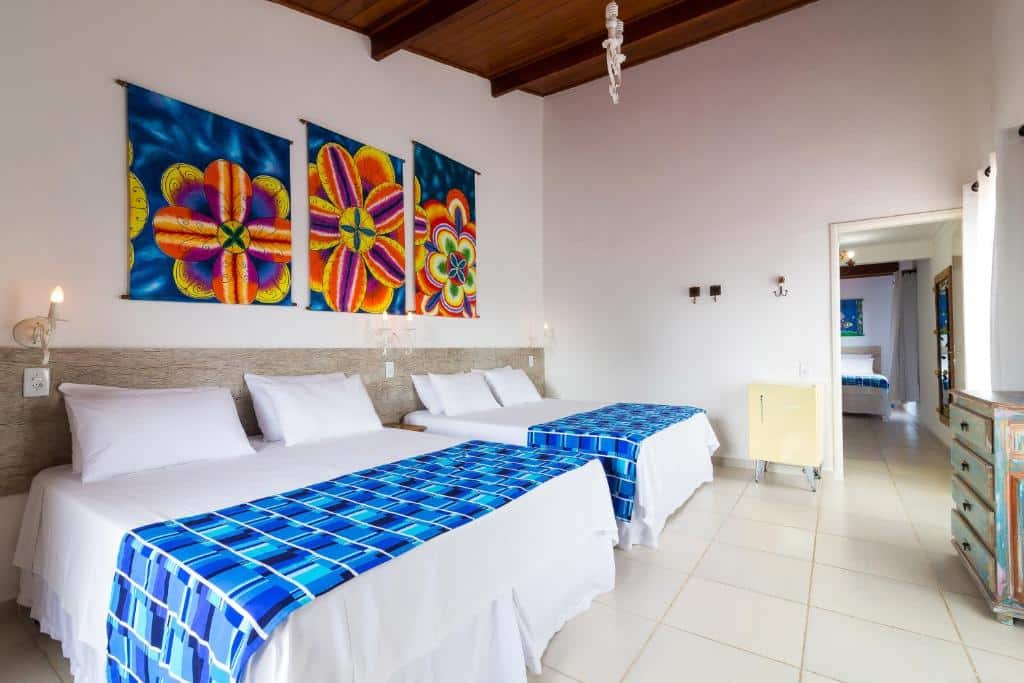 Quarto duplex em Hotel Village Enseada, duas camas de casal com lençóis brancos e azul, uma decoração clara, com alguns quadros coloridos, espaço amplo, um frigobar, uma janela e uma cômoda