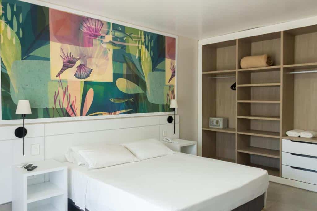 Quarto em Vistabela Resort & Spa, uma cama de casal, um armário aberto do lado direito, um mural com desenhos tropicais sob a cama, tudo branco e dois abajures, para representar pousadas em São Sebastião