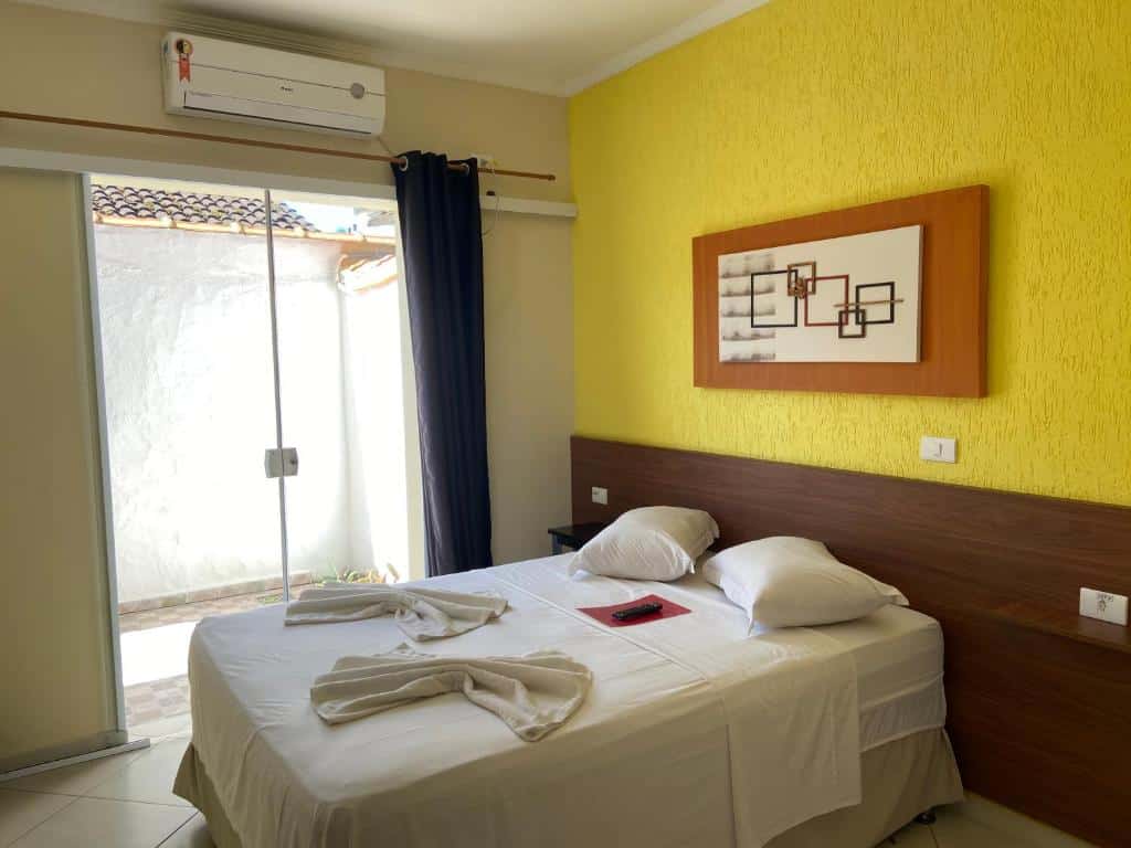 Quarto Werneck Residence Suites, uma cama de casal com travesseiros e toalhas sob a cama, uma parede amarela, do lado esquerdo uma sacada, um quadro sob a cama e um ar-condicionado