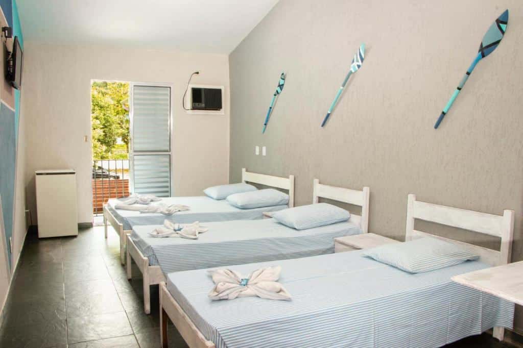 Quarto no Hotel Costa Azul, uma cama de casal e duas de solteiro, uma sacada, ar-condicionado, televisão, uma frigobar e alguns enfeites rústicos nas paredes, para representar pousadas na Praia da Enseada em Ubatuba