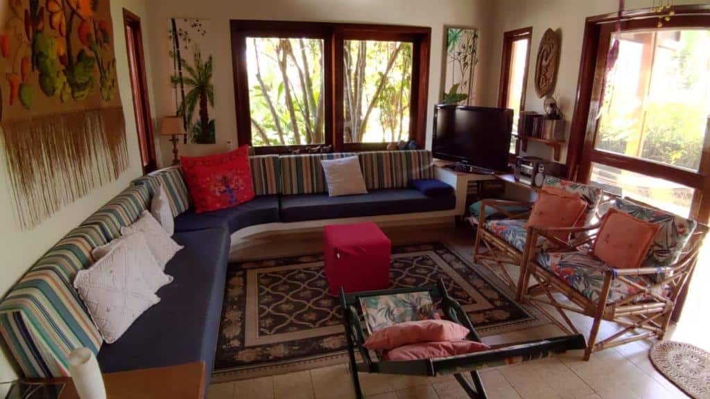 Sala de estar da Aconchego na Praia, dois sofás que formam um L, algumas almofadas, duas cadeiras, um tapete, uma televisão, alguns enfeites rústicos e duas janelas amplas