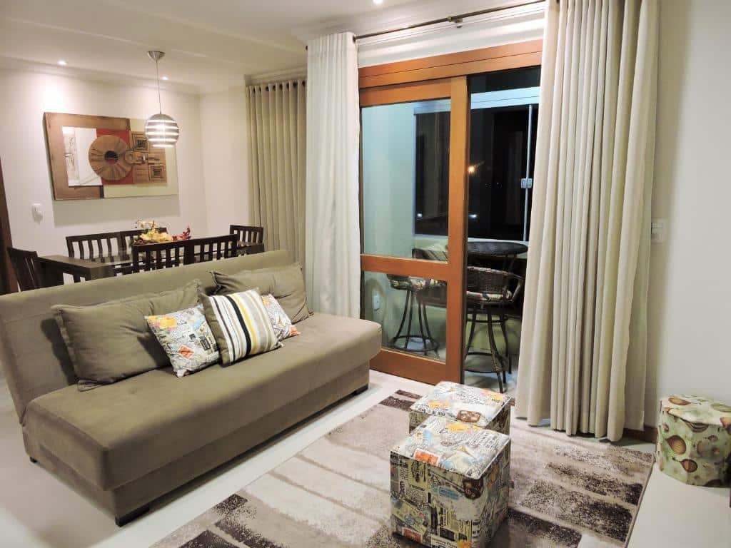 Sala de estar do Apto no centro de Gramado, uma sacada com cortina e local para sentar, um sofá e alguns bufes, além da mesa de jantar com seis lugares e um pequeno lustre