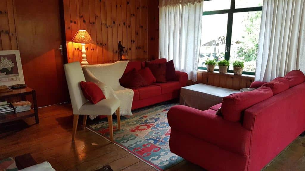 Sala da Casa Sirin, dois sofás, uma janela ampla, uma poltrona, um abajur, tudo em madeira e nas cores branco e vermelho, para representar airbnb em Campos do Jordão