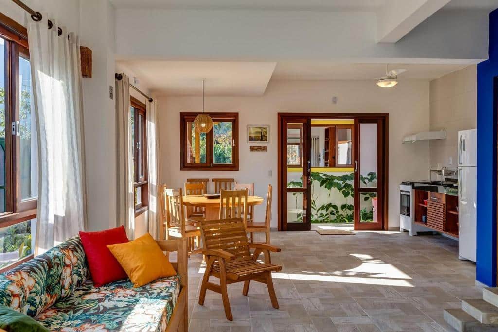 Sala de estar e jantar em Vila dos Manacás, um local amplo e arejado com muitas janelas e uma porta de vidro, a cozinha está do lado direito, do esquerdo, tem algumas cadeiras e mesa de madeira, além de um sofá
