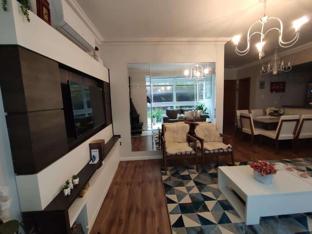 Sala de estar e jantar no Gramado Garden Centro, uma mesa com seis lugares, uma varanda ampla, duas poltronas, um tapete geométrico e uma televisão, para representar airbnb em Gramado